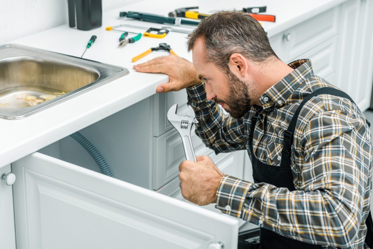 Man in plaid shirt and overalls fixes a pipe under a kitchen sink using a wrench. Tools are scattered on the counter, suggesting repair work.