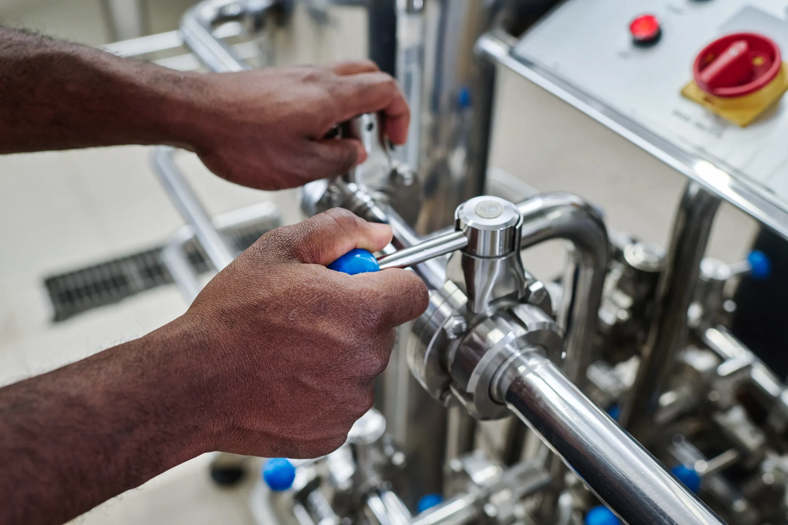 Hands adjust a wrench on a stainless steel pipe in an industrial setting. The background includes control knobs, indicating a mechanical or engineering task.