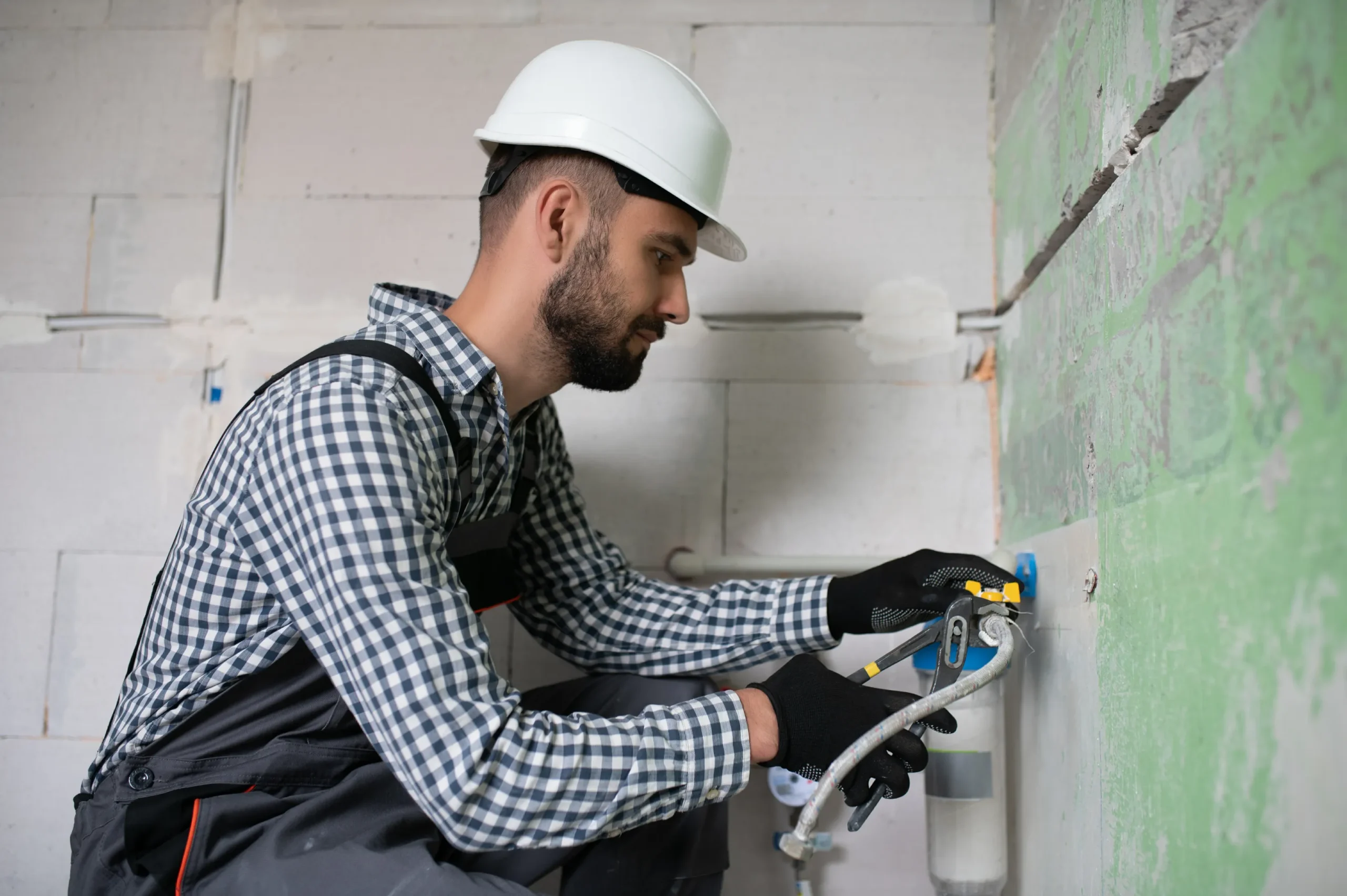 Man in a checkered shirt and hard hat using a spray gun on a green wall. He wears black gloves, conveying focus and precision in the construction work.