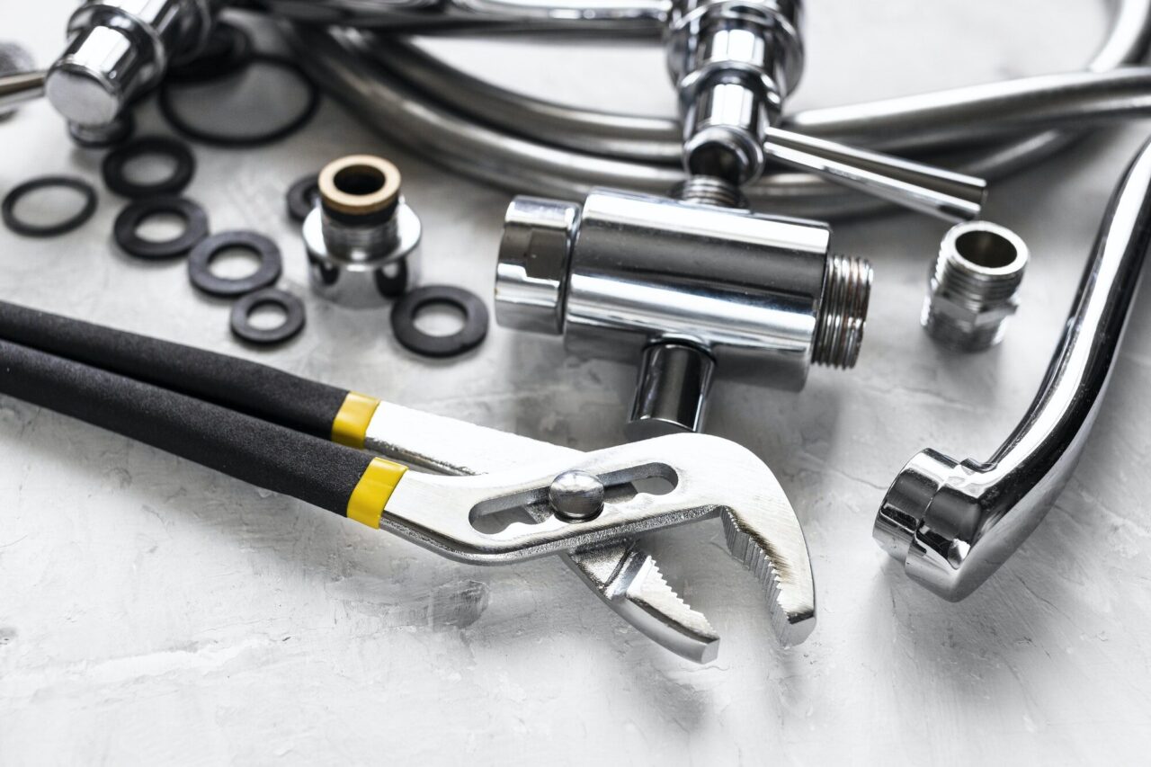 Alt text: "Plumbing tools and parts, including an adjustable wrench, metal pipes, and rubber washers, on a light surface, conveying a sense of preparation for repair work."